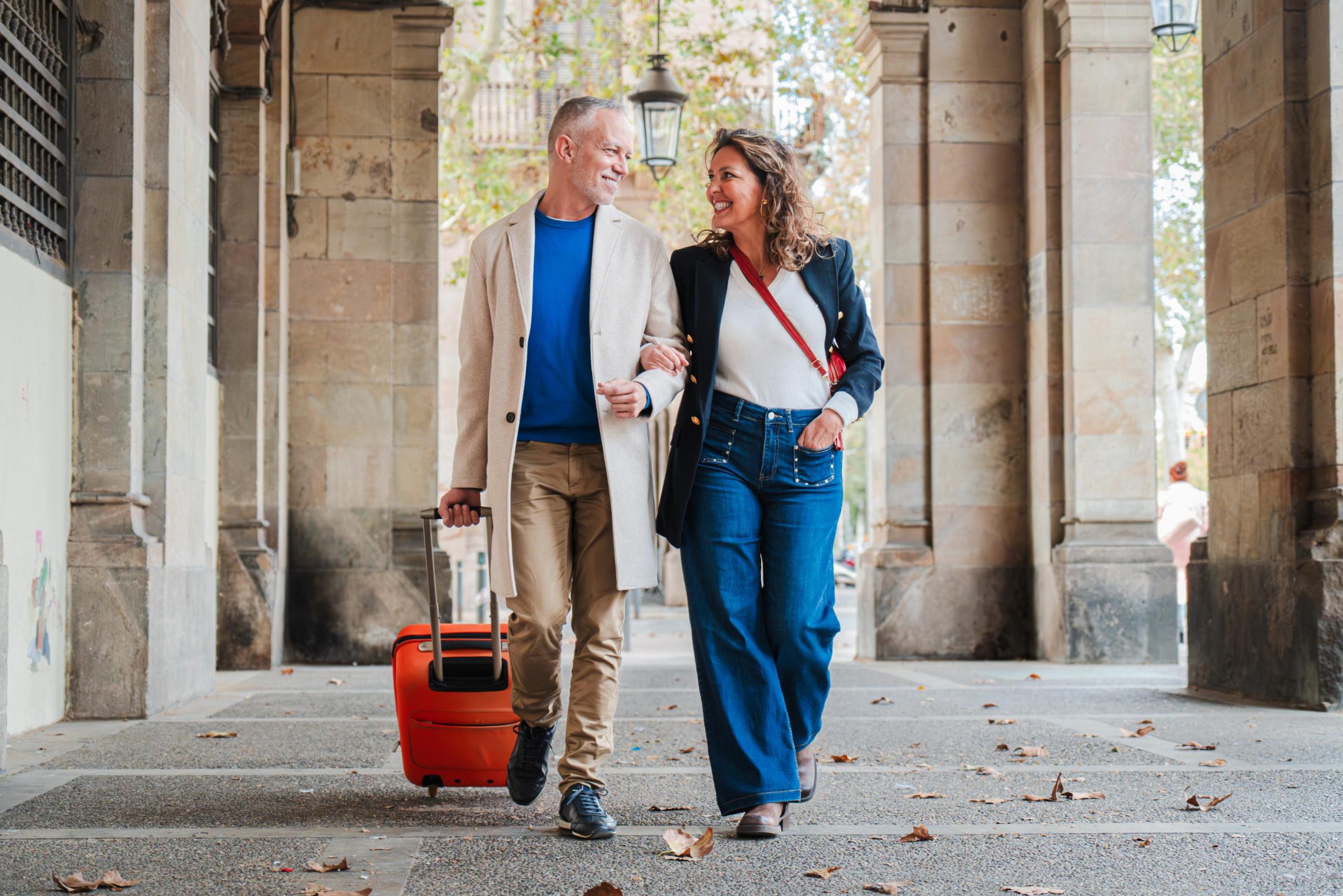 Pareja madura paseando felizmente mientras disfrutan de sus vacaciones en España, llevando una maleta naranja por una calle con arcadas.